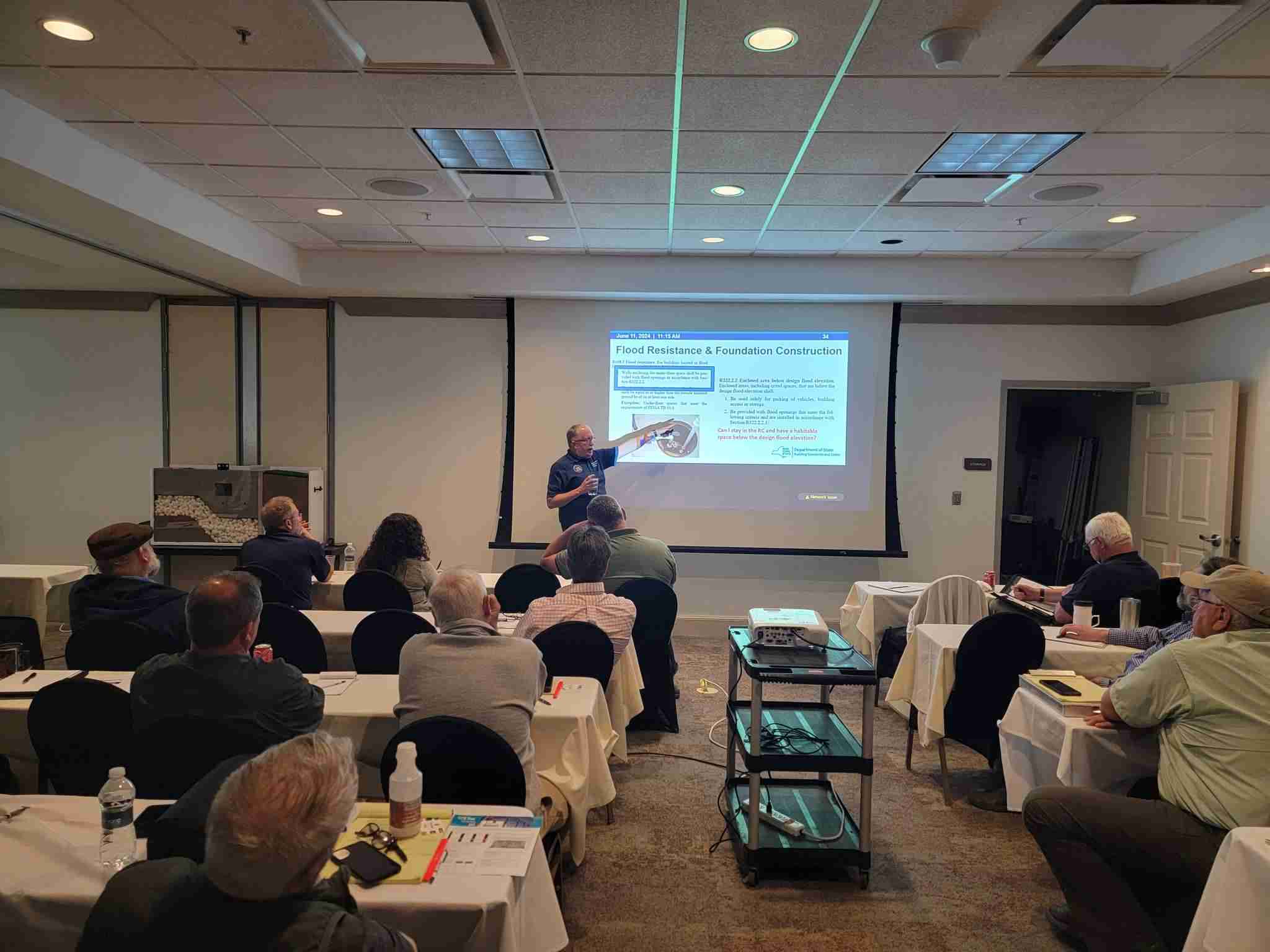 480533611_1020476973443462_9001781734809003668_n A presenter stands at the front of a classroom, speaking to seated attendees while displaying a slide about flood resistance and foundation construction on a projector screen.