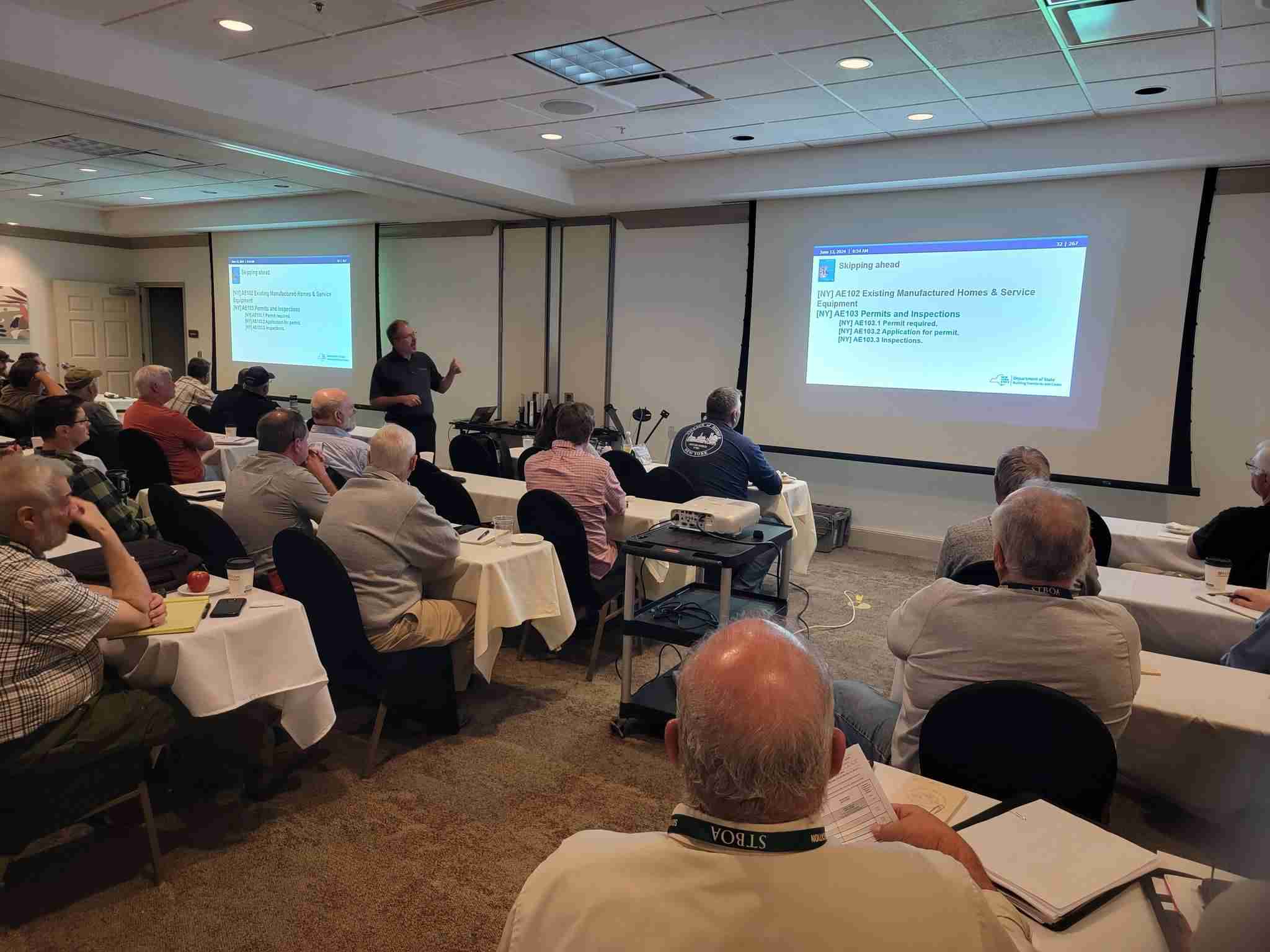 A group of people sit in a conference room facing a presenter and a projected slide titled “2023 Existing Manufactured Homes & Service.” The audience appears attentive, taking notes during the presentation.